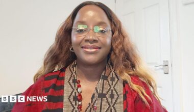 Bami is wearing a red and black dress, which has black and cream patterned detailing around the neck-line. She has long, wavy light brown hair and is wearing frameless glasses. She is sat on a grey seat with a white wall and door behind her.