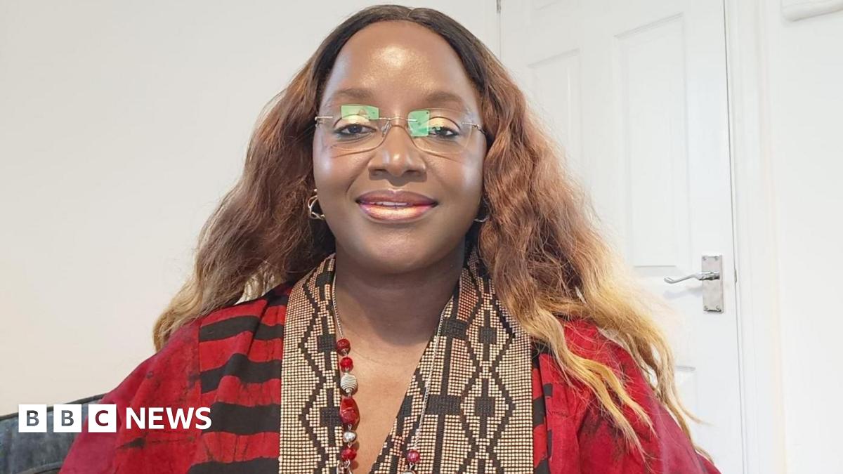Bami is wearing a red and black dress, which has black and cream patterned detailing around the neck-line. She has long, wavy light brown hair and is wearing frameless glasses. She is sat on a grey seat with a white wall and door behind her.