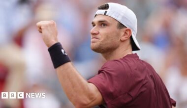 A man on a tennis court raising a clenched fist. He is wearing a baseball cap and maroon t-shirt.