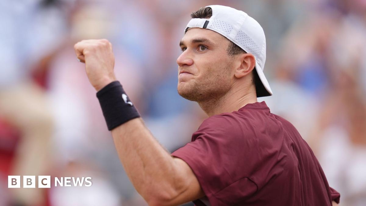 A man on a tennis court raising a clenched fist. He is wearing a baseball cap and maroon t-shirt.