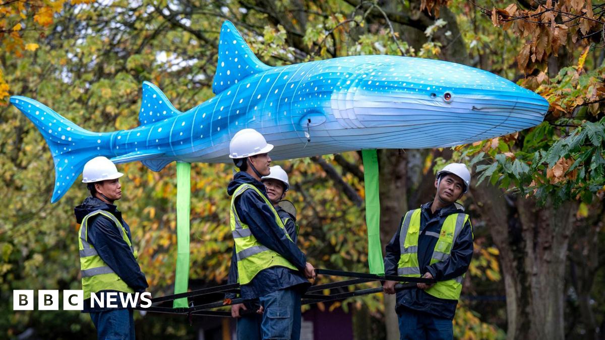 Four men in hard hats and fluorescent jackets carry a lantern shaped like a blue whale above them. Trees can be seen in the background with green and orange leaves
