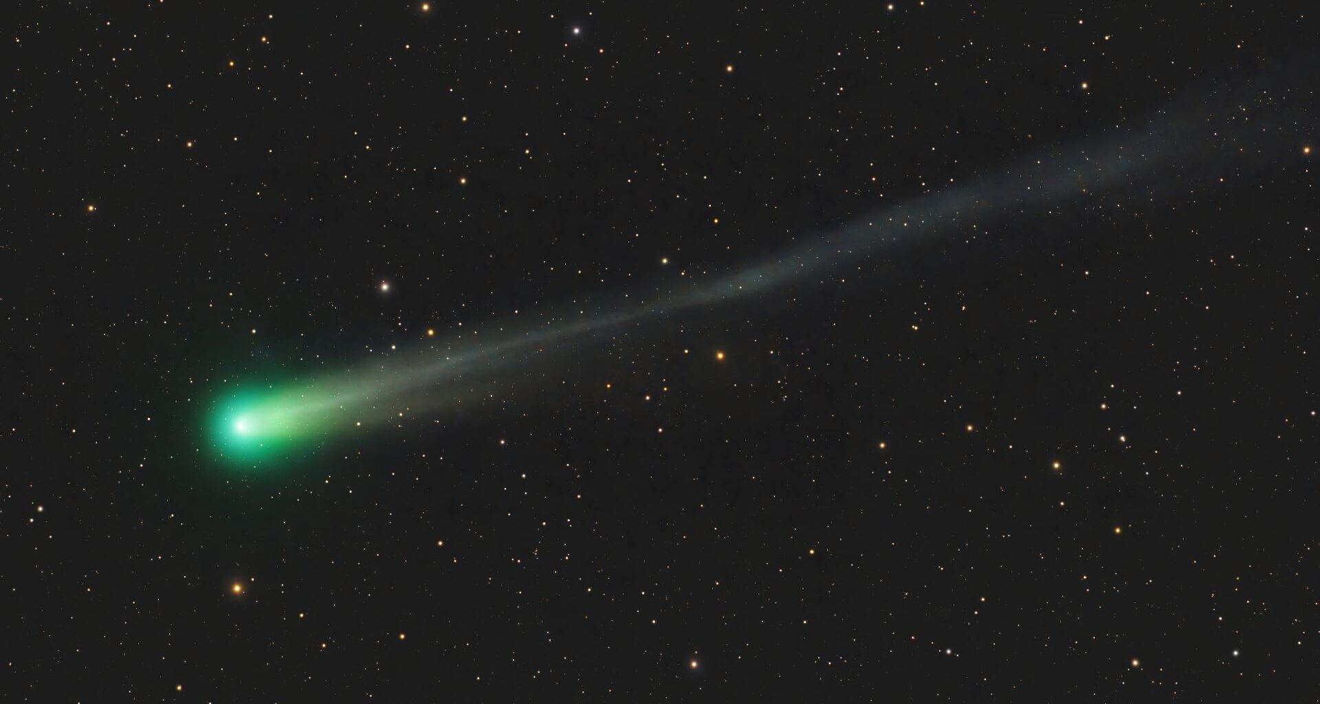 A comet with a green corona and long tenuous tail is pictured streaking left to right across a black sky peppered with stars.