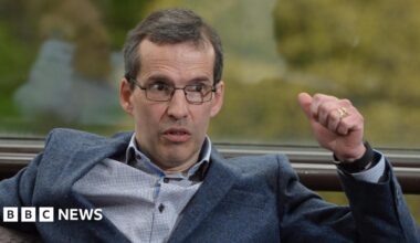 Mr Paisley wears black rectangular glasses and has short grey hair. He speaks to the right of the camera shot as he wears a dark suit jacket and a white shirt. He has brown eyes