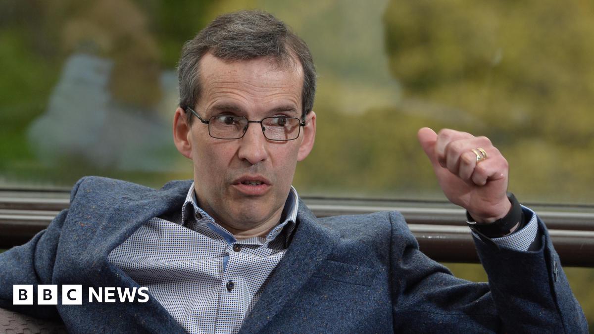 Mr Paisley wears black rectangular glasses and has short grey hair. He speaks to the right of the camera shot as he wears a dark suit jacket and a white shirt. He has brown eyes