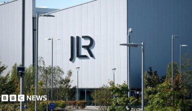 A view of a large, grey factory building sitting behind a parking lot lined with trees and shrubs. On the side of the building are large letters spelling JLR.