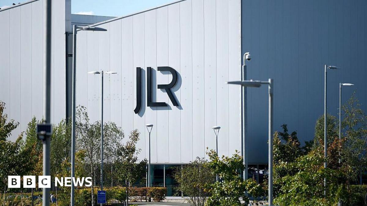 A view of a large, grey factory building sitting behind a parking lot lined with trees and shrubs. On the side of the building are large letters spelling JLR.