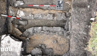 A small square burial site is a mixture of brown mud and grey stone. There are two red and white thin sticks lying horizontally and parallel to each other.