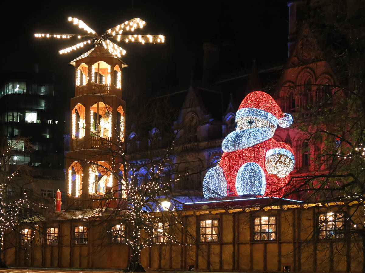 big santa sat in albert square, manchester next to christmas-markets stalls and lit-up windmill
