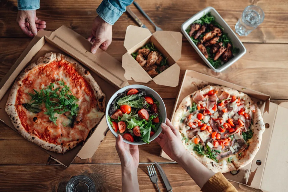 Hands reaching for a spread of pizza, salad, and chicken wings on a wooden table. Pizza boxes are open, and a salad is in a bowl