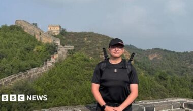 Will Blisset wears a black t-shirt, shorts and cap. He stands in front of a section of the Great Wall of China. You can see lush green trees in the background and a  walk that snakes up into the hills.