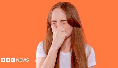 A woman with ginger hair and wearing a white T-shirt sneezing while pinching her nose, against a bright orange background