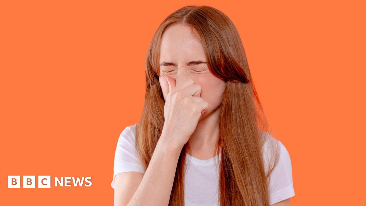 A woman with ginger hair and wearing a white T-shirt sneezing while pinching her nose, against a bright orange background