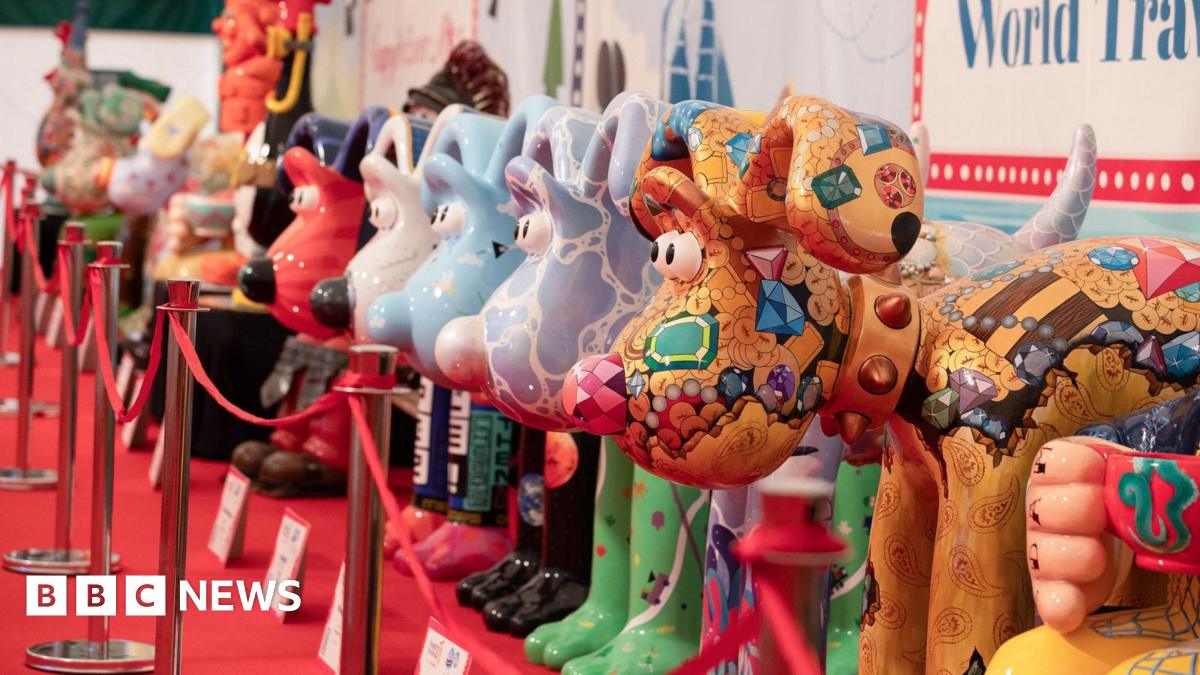 Large statues of Aardman's Gromit character are lined up on a red carpet, positioned behind red tape. They are all painted with colourful designs, with the closest one a gold colour with gem stone paintings all over its face and body.