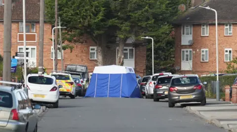 George Tryfonidis/BBC A blue and white forensics tent stands in the middle of a residential street with cars parked on either side of the road. A police car is next to the tent