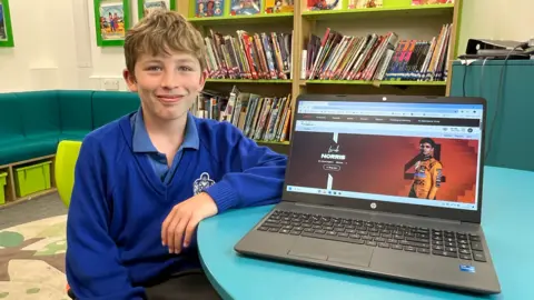 A smiling young boy in blue school uniform sitting while resting his left arm on a table. On the table is an open laptop screen with an image of Lando Norris on it