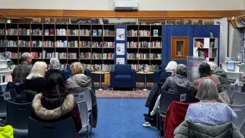 A wall filled with books can be seen, a blue arm chair sat in front. Rows of people sitting facing the wall of books.