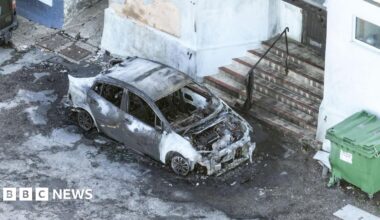 A scorched car outside the mosque. It is parked in front of the entrance steps, which are covered in black soot and have partially melted railings.