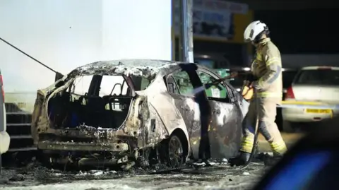 Eddie Mitchell A car is scorched as a firefighter sprays it with water. 
