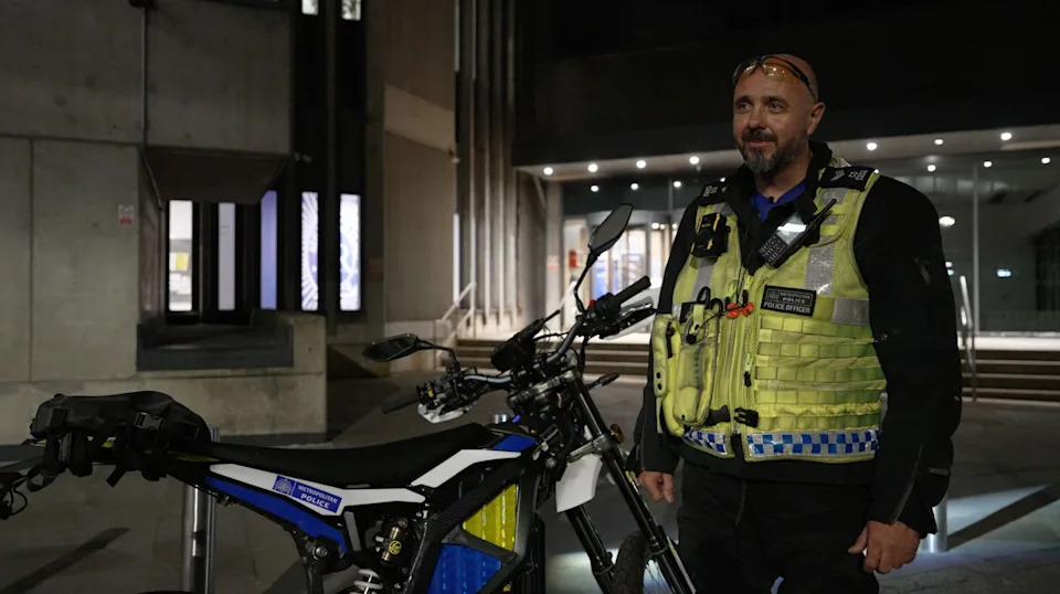 Police officer stands next to one of the force's own new high-powered electric bike. The officer is wearing a high vis police gilet with phone attached, and various equipment. In the background is a concrete facade of police building that is lit up in the windows. The officer has goggles resting on his forehead, a dark brown beard and shaven head.