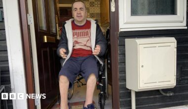 A man has dark, close-shaven hair and is wearing a grey hoodie, a red T-shirt and blue shorts. He is using a wheelchair and is in the doorway of a flat with a step outside.