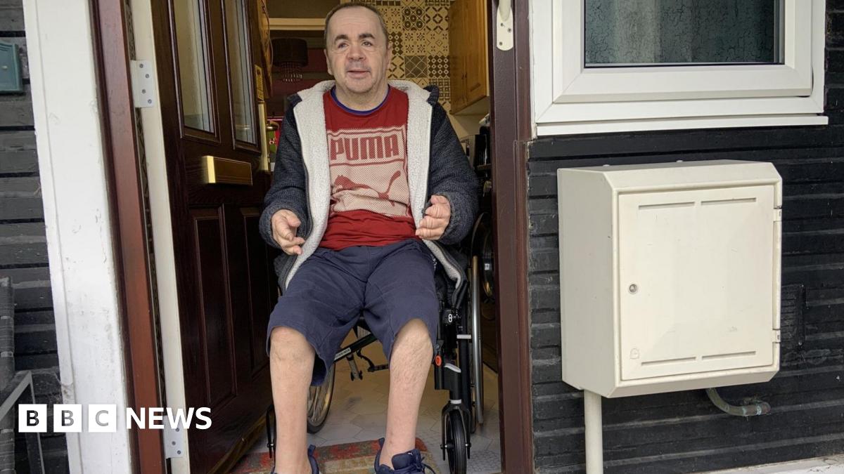 A man has dark, close-shaven hair and is wearing a grey hoodie, a red T-shirt and blue shorts. He is using a wheelchair and is in the doorway of a flat with a step outside.