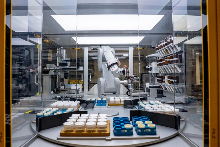 A large white robotic arm holding a glass vial inside a glass box in a lab with many other containers in racks.