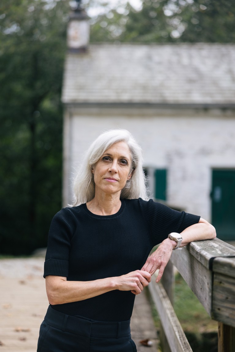 Susan Monarez poses for a portrait leaning on a wooden bridge with an old stone building behind her