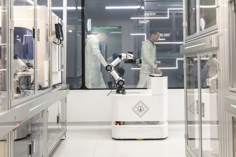 A robot is seen in a bright lab while two people walk past behind a window in the background