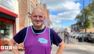 Nigel Beecroft has short grey hair and wears a purple running vest over a T-shirt. He is standing on a high street with shop fronts and parked cars behind him.