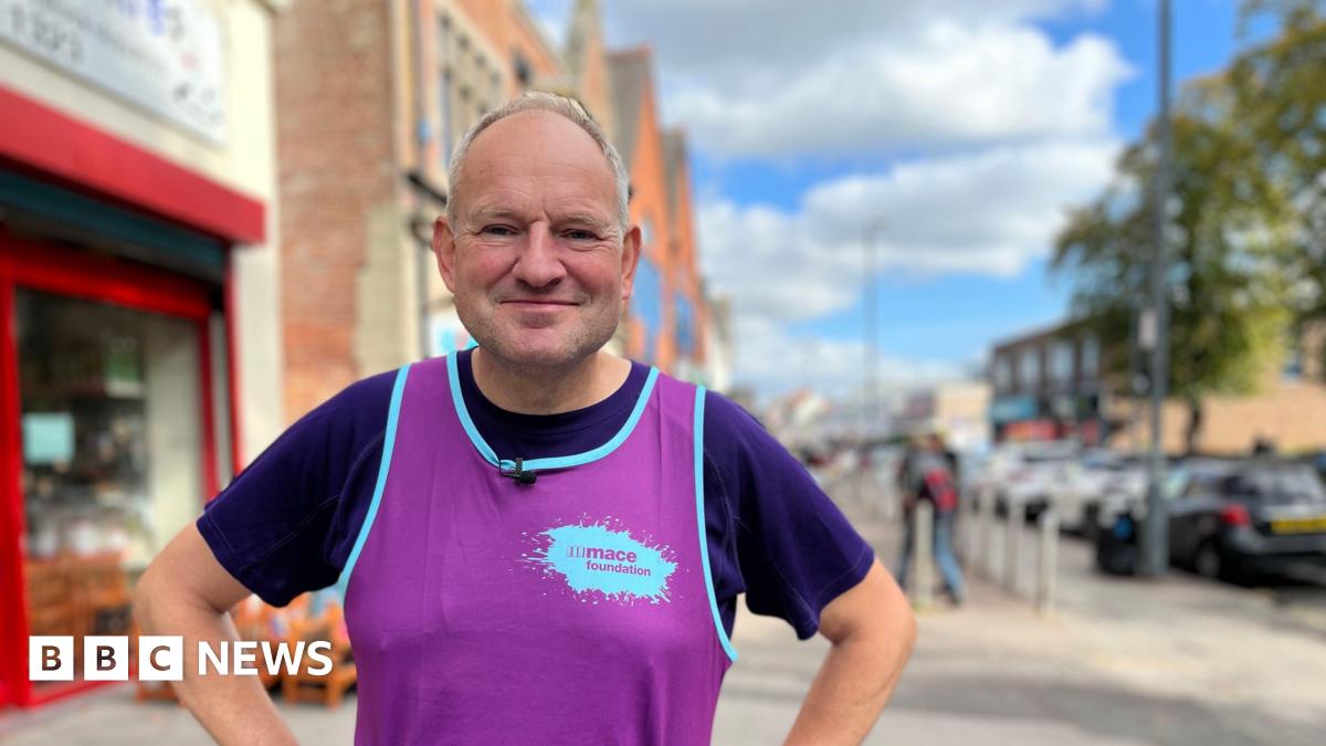 Nigel Beecroft has short grey hair and wears a purple running vest over a T-shirt. He is standing on a high street with shop fronts and parked cars behind him.