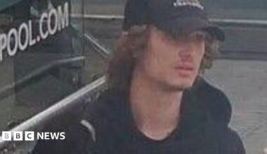 A CCTV image showing a young man with brown curly hair emerging from under a black baseball cap. He is wearing a black Monterrain hoodie