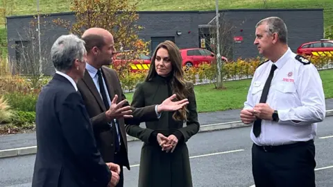 Prince William and his wife Catherine stand talking to two men outside a fire service training college building. William is wearing a brown blazer over a light shirt and dark tie.  He is gesticulating with his hands as he talks.  Catherine is wearing a long, dark olive green coat, buttoned up to the neck.   A firefighter wearing a white uniform shirt and black tie is listening to the prince.  Another grey-haired man in a navy suit has his back to the camera. 