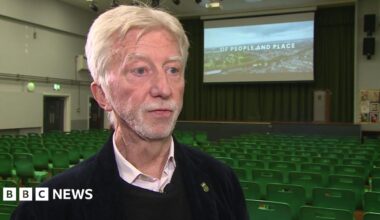 Jimmy Mulville, who has grey, parted hair and is wearing a black jumper with a pink, open-collared shirt underneath, is standing in a school assembly hall. There are rows of empty green seats behind him along with a large screen with the opening title of his documentary superimposed over an aerial shot of Walton.