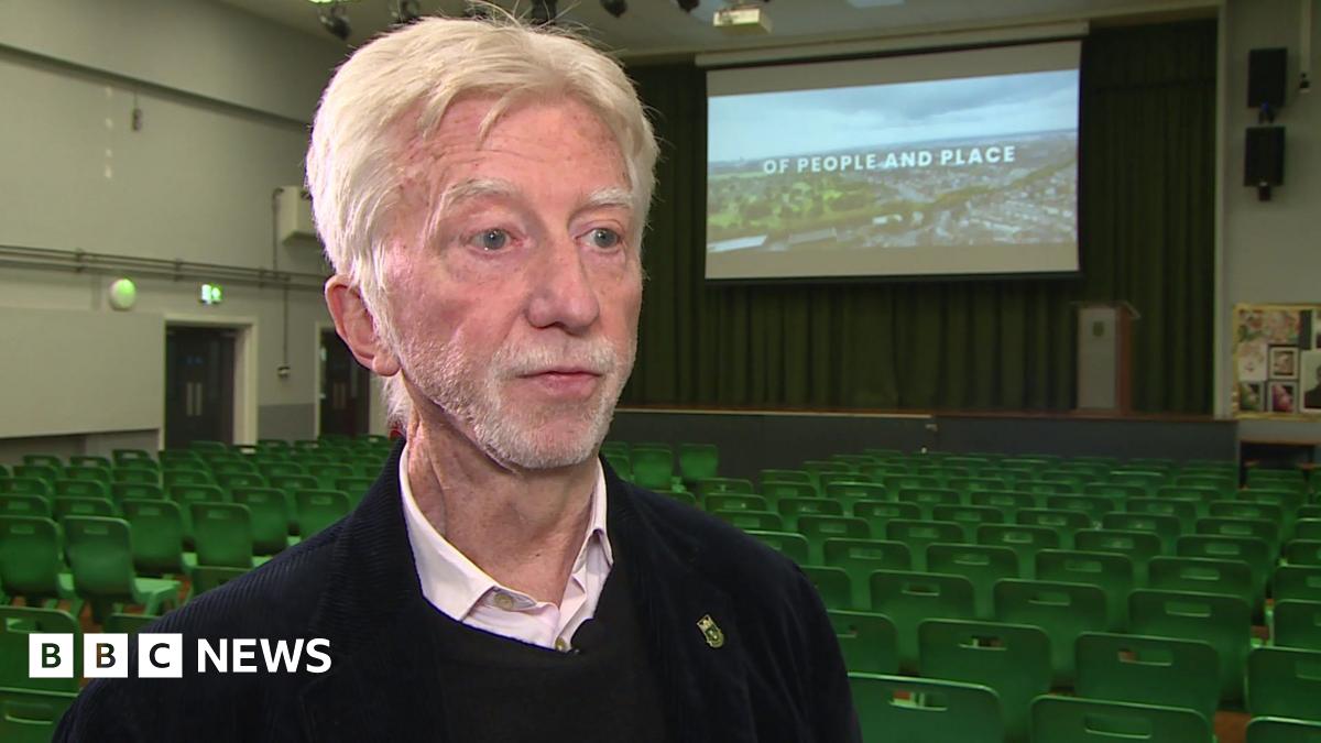 Jimmy Mulville, who has grey, parted hair and is wearing a black jumper with a pink, open-collared shirt underneath, is standing in a school assembly hall. There are rows of empty green seats behind him along with a large screen with the opening title of his documentary superimposed over an aerial shot of Walton.