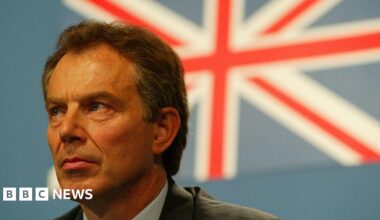 Close-up of Tony Blair at the G8 Summit, looking slightly to the left with a serious expression. He is wearing formal attire, and behind him is a large, blurred Union Jack flag on a blue background.