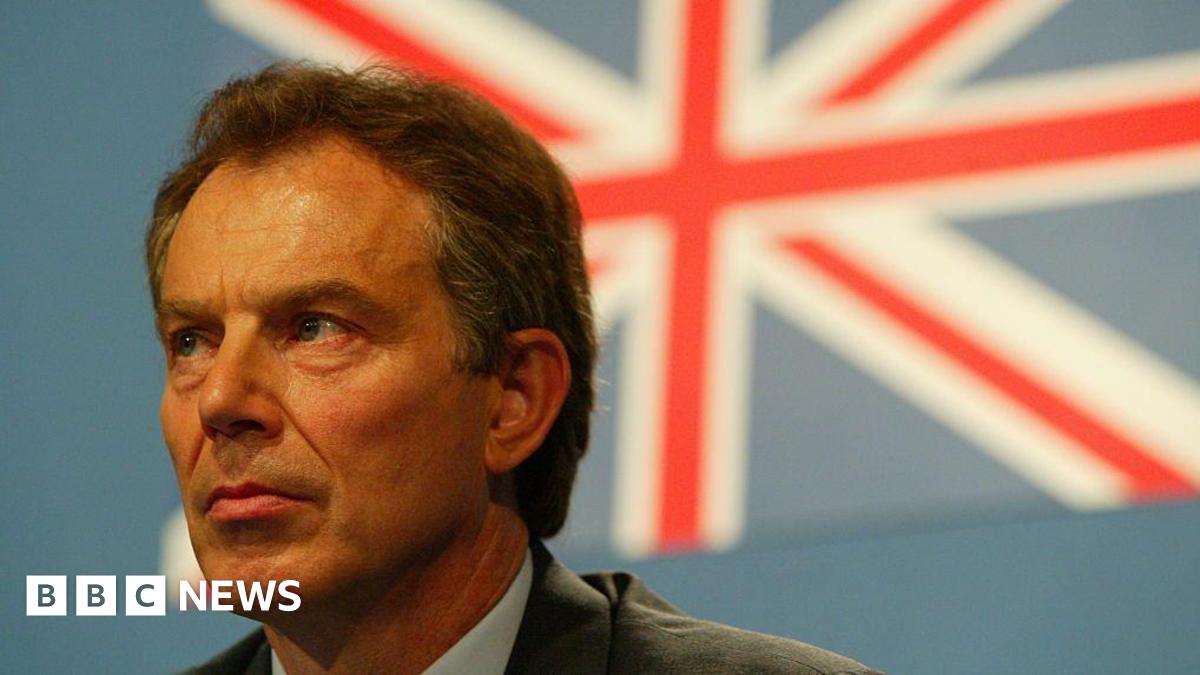 Close-up of Tony Blair at the G8 Summit, looking slightly to the left with a serious expression. He is wearing formal attire, and behind him is a large, blurred Union Jack flag on a blue background.