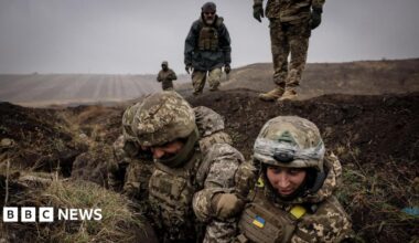 Ukrainian soldiers, in camouflage military clothing, stand in a trench during a training exercise. Behind them, more personnel are standing in an otherwise empty, brown field.