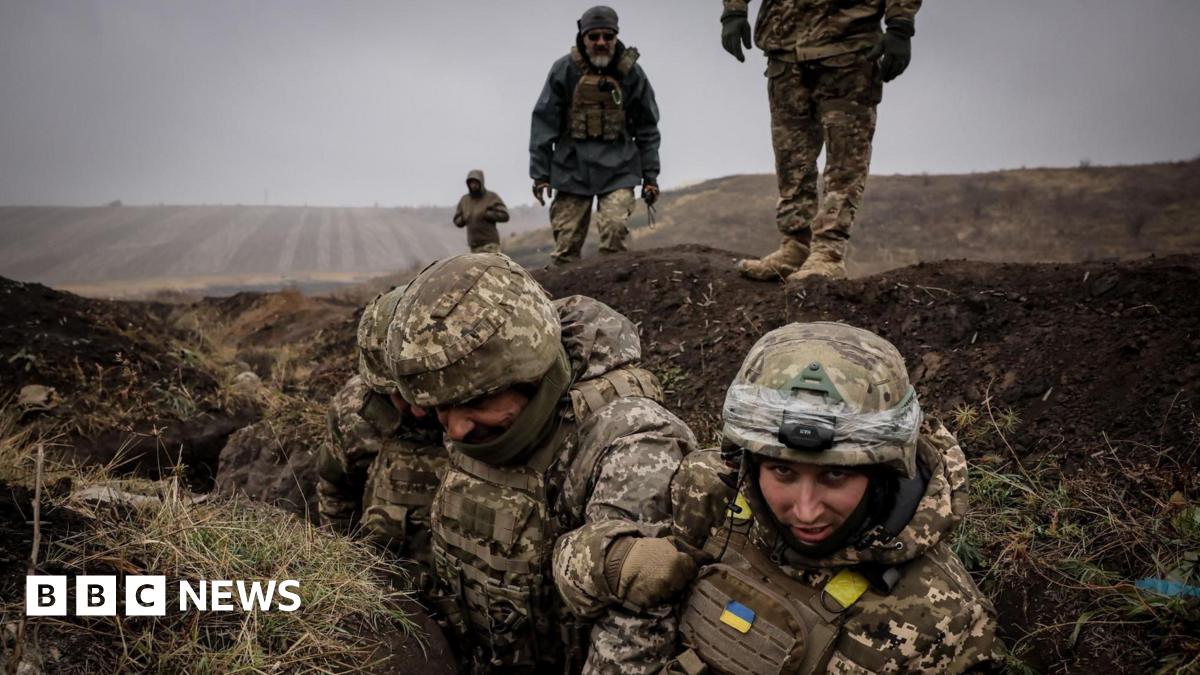Ukrainian soldiers, in camouflage military clothing, stand in a trench during a training exercise. Behind them, more personnel are standing in an otherwise empty, brown field.