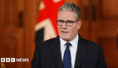 Sir Keir Starmer speaks in a wood-walled room in front of two union jack flags. He is wearing a black suit jacket, a blue tie with white polka dots, and a white shirt. He has a serious expression.