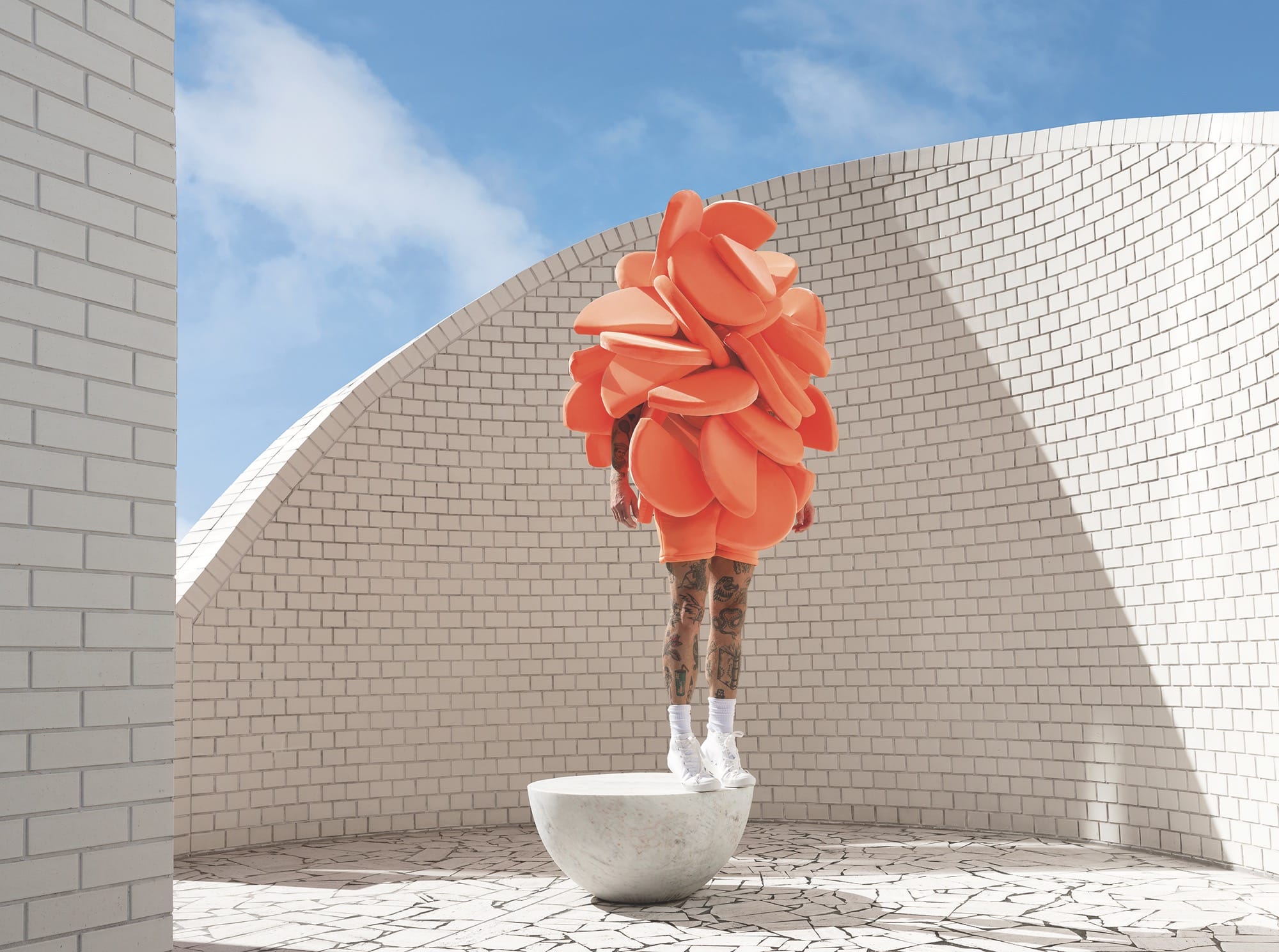 a photo by Gerwyn Davies of a figure in a bright orange abstract costume, standing amid contemporary architecture
