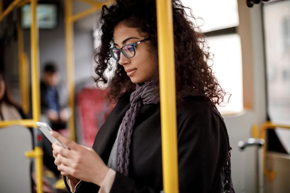 A person with curly hair and glasses stands on a bus, looking at their smartphone. They are wearing a scarf and coat