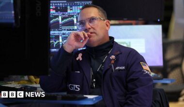 A trader in a dark blue coat with an American flag on his sleeve sits near computers at the New York Stock Exchange in October 2025, his eyes lifted to the screens above him.
