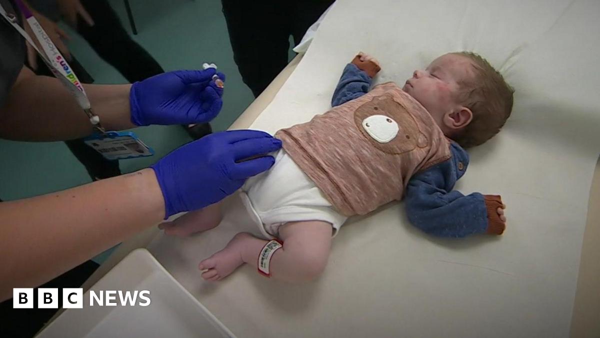 Baby Harry lying on a bed wearing a white babygrow and a brown and blue top with a teddy bear's face on the front. Harry has his eyes closed and the hands of a nurse wearing blue surgical gloves holding a needle about to administer the injection.