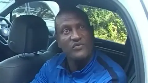 PA Media Hadush Kebatu sits in the back of a car. He wears a bright blue zip-up top with a black stripe down the shoulder. The car is parked and he is the only passenger, sat in the back seat with the door open. He is speaking to someone off camera. 