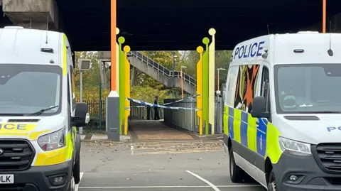 Two police vans outside a station. The white vans have blue and white markings and the word 'police' on. There is blue and white police tape at the station entrance. 