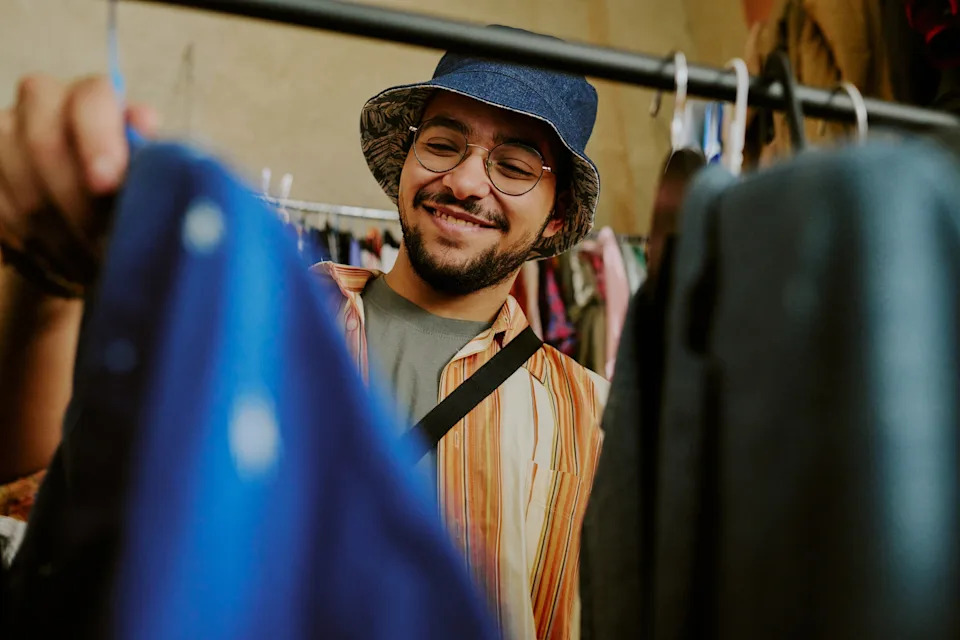 Man in a striped shirt and blue hat smiles while browsing clothes on a rack