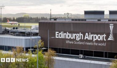 The exterior of Edinburgh Airport with an Easyjet flight landing on the runway in the top left hand corner of the shot