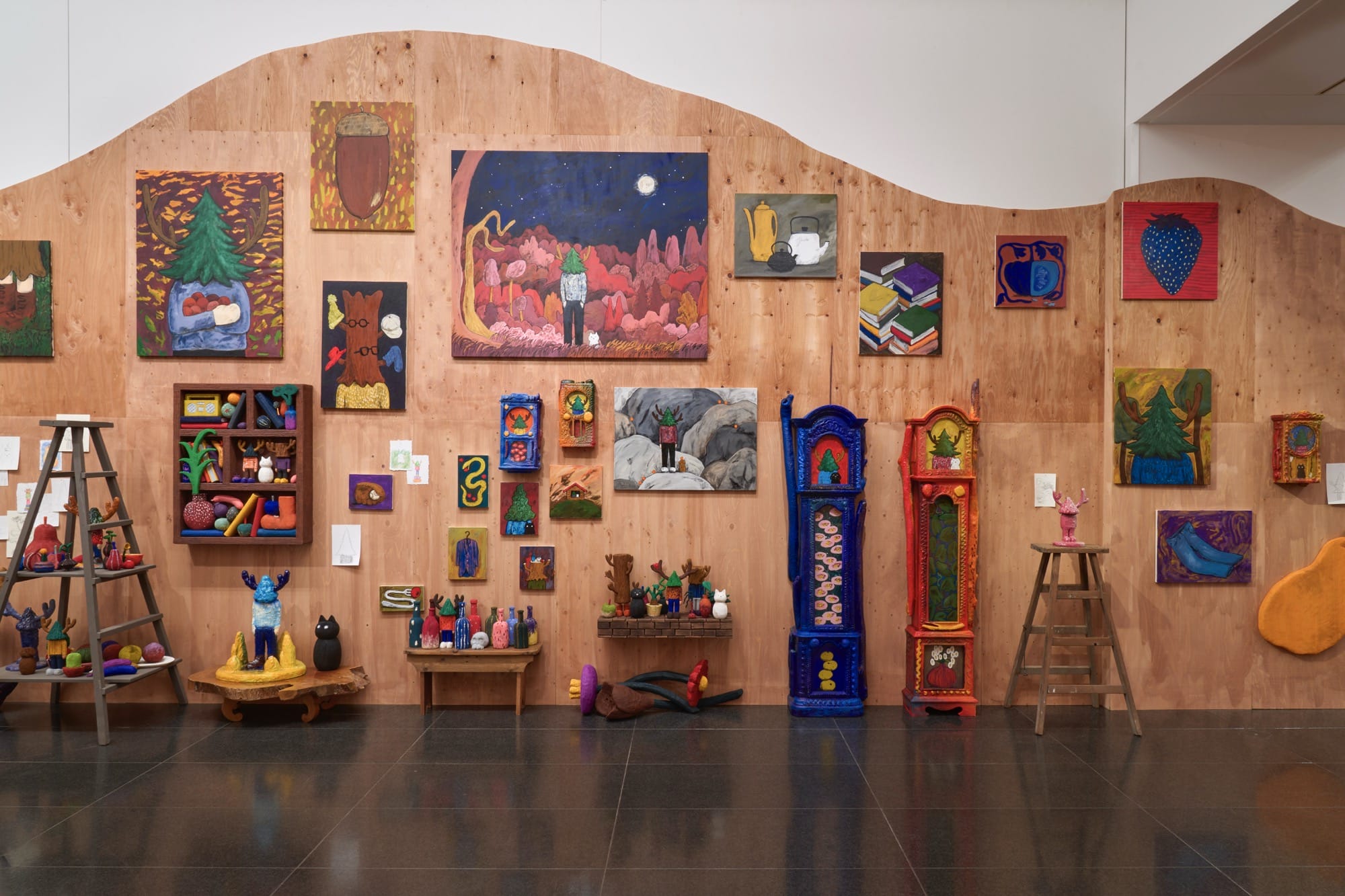 a large-scall wall installation by Yuichi Hirako with a curving background of plywood, with a salon-style hang of paintings and sculptures