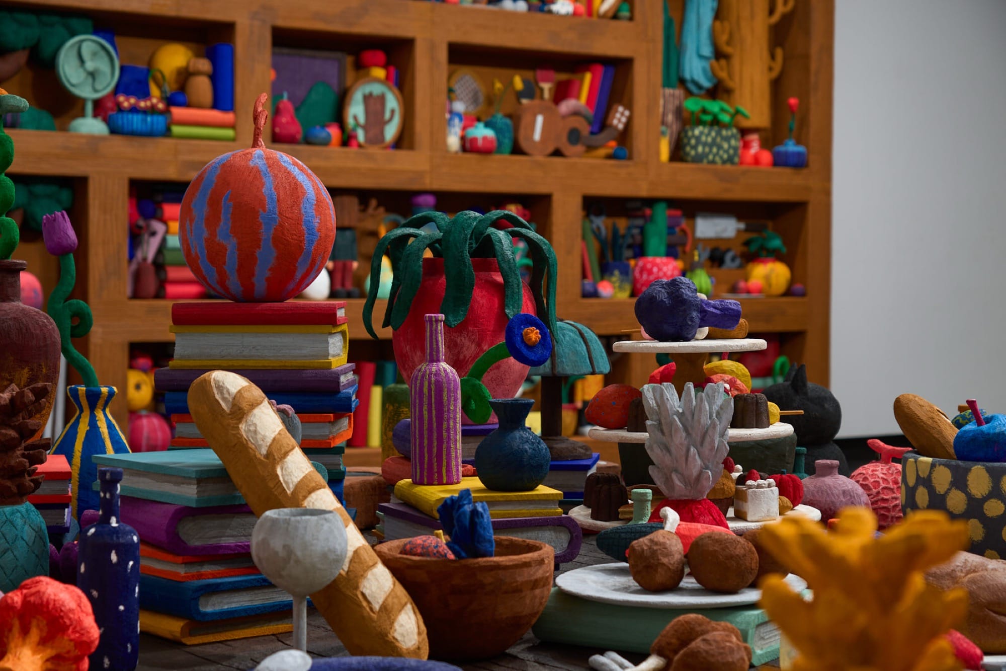 a detail of a sculptural installation by Yuichi Hirako of a large table and an oversized shelf covered with cartoonish domestic forms like vases, food, candlesticks, and other objects
