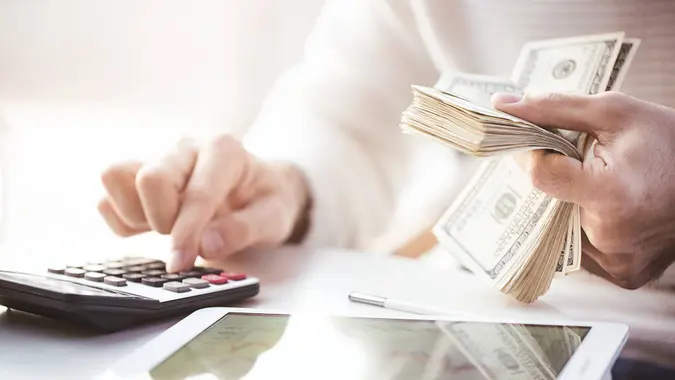 A close-up of two hands, one using a calculator and the other holding a stack of money.